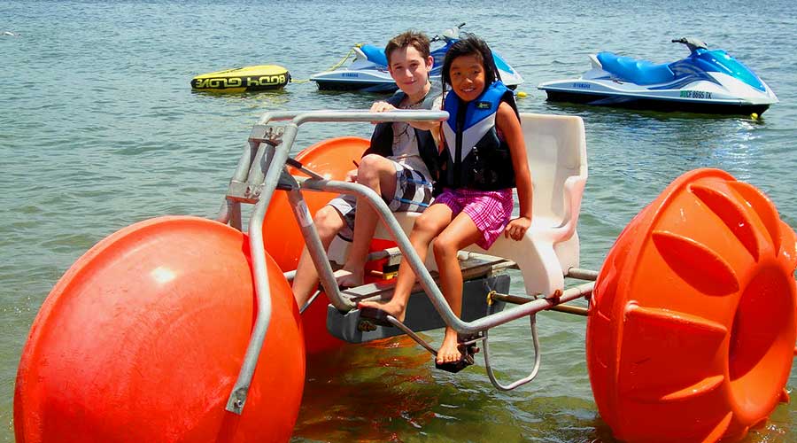 Two kids having fun on a water tricycle at a hotel resort Orange Aqua-Cycle™ Water Trike at Campland on the Bay in San Diego for their recreation water equipment for a rental business